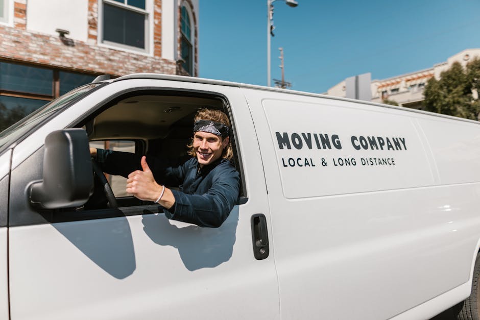 A young man with shoulder-length hair, wearing a dark headband and a navy blue jacket, is sitting in the driver's seat of a white moving van with the text 'MOVING COMPANY, LOCAL & LONG DISTANCE' print