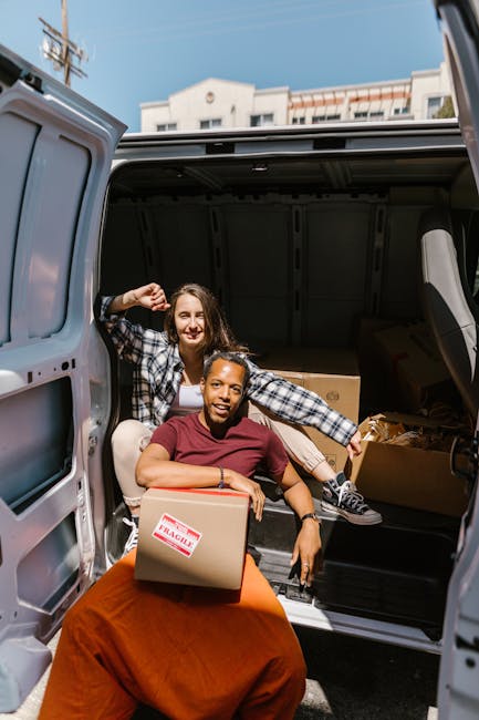 A young man with shoulder-length hair, wearing a dark headband and a navy blue jacket, is sitting in the driver's seat of a white moving van with the text 'MOVING COMPANY, LOCAL & LONG DISTANCE' print