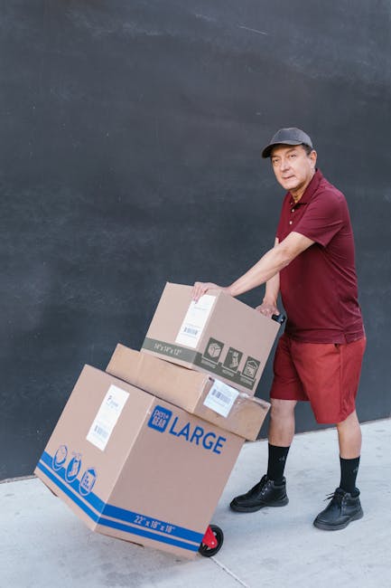 A man wearing a maroon polo shirt, matching shorts, black socks, and black shoes is seen pushing a red-handled hand truck loaded with three cardboard boxes of varying sizes outside a building with a dark grey wall. The boxes are sealed with packing tape, and some have labels indicating contents or sizes, including one marked 'LARGE'. The largest box is positioned at the bottom of the hand truck, supporting the smaller boxes stacked atop. The scene appears to be part of a home relocation or furniture transport process, with the man preparing to load or unload the boxes as part of a professional removals service offered by Man with Van Waddon, which specialises in removals and moving logistics in the Waddon area. The environment is lit with natural light, the surface beneath the boxes is concrete or similar pavement, and the overall setting reflects an organized loading or packing activity essential to household moves or same-day emergency relocations.