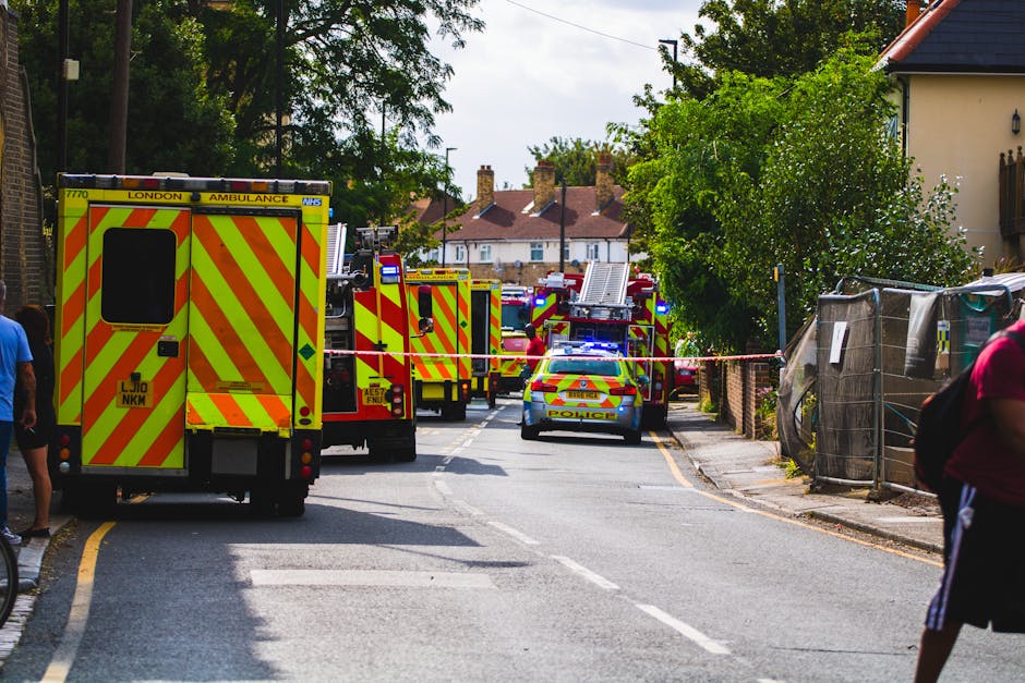 A street scene during an emergency response shows multiple fire trucks and a police car blocking the road, with emergency lights flashing. Firefighters are visible near the vehicles, possibly preparing equipment for a home rescue or safety operation. The street is lined with residential houses, some with pitched roofs and chimneys, and has trees providing greenery along the pavement. A few pedestrians stand nearby, observing the scene. The road is partially cordoned off with red and white tape, and some household items or construction materials are visible on the sidewalk. Inside a property, additional emergency and rescue equipment may be present, indicating a coordinated response to an incident. This image reflects the logistical complexity of emergency moves or safety measures in residential areas, with various vehicles and personnel engaged in their tasks. Occasionally, Man with Van Waddon might support such relocation or emergency transfer services, ensuring safe and efficient household or furniture transport amidst urgent situations.