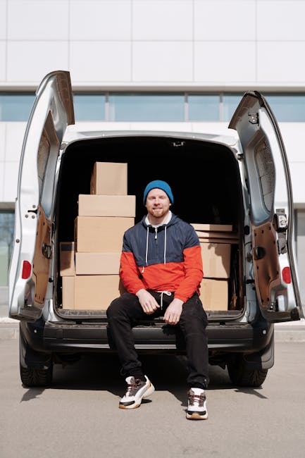 A man dressed in a blue beanie, navy jacket with red and white accents, black trousers, and white sneakers is sitting on the edge of an open van’s loading area. The van, parked on a paved surface in front of a large modern building with white tiled exterior, contains several neatly stacked cardboard boxes of various sizes, some wrapped in plastic for protection. The interior of the van appears to have a dark, spacious cargo area ready for furniture transport and moving supplies. The man is smiling and facing forward. The scene illustrates a home relocation or furniture transport process, with visible packaging materials and the vehicle positioned for loading or unloading. Man with Van Waddon specializes in removals services, assisting in packing and moving logistics.
