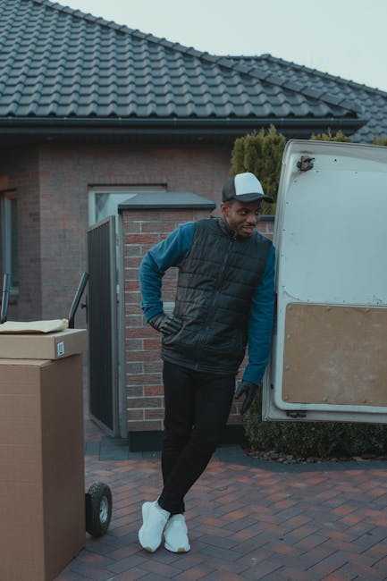 A man standing in front of an open van with its rear doors swung wide open, revealing several cardboard boxes neatly stacked inside for a home relocation or furniture transport. The man is dressed in a red and navy blue jacket, black trousers, and a blue beanie, with arms crossed and a neutral expression. The scene is set outdoors in a paved area in front of a modern, white multi-storey building with large windows, suggesting a suburban or urban environment. Visible inside the van are packing materials and boxes prepared for moving, indicative of the loading process involved in professional removals and packing and moving services. The van is parked on a level surface, and the environmental lighting is bright, highlighting the details of the boxes, the vehicle, and the person's attire. This image illustrates the process of loading goods for a local or long-distance move, emphasising logistics and transportation elements relevant to house removals by Man with Van Waddon.