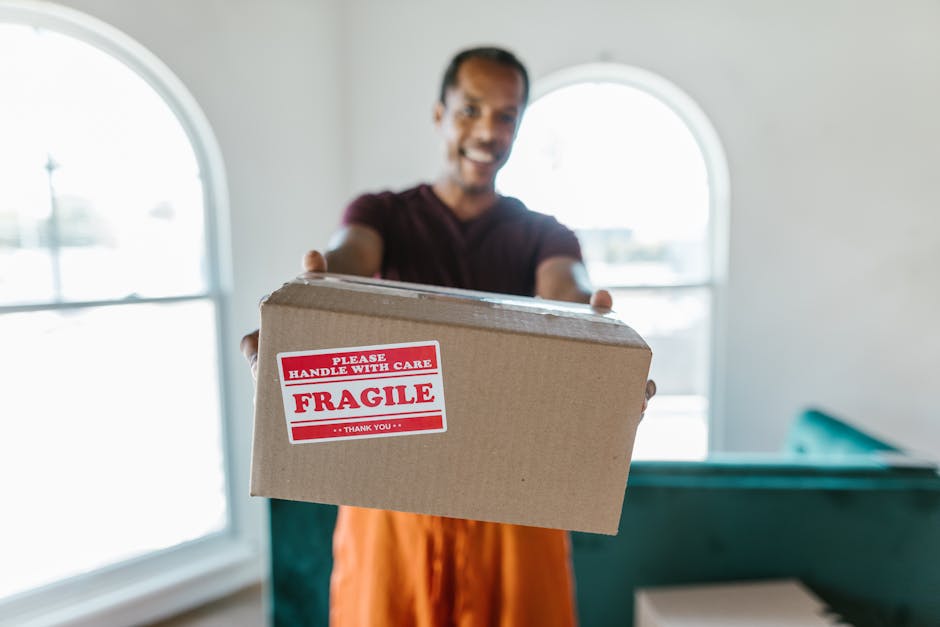A man with dark, curly hair and a beard is kneeling on a wooden floor inside a room, surrounded by stacked cardboard moving boxes of various sizes, some labeled for bedrooms and kitchen. He is wearing