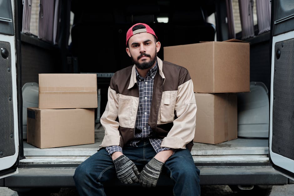 A young man with a beard, wearing a red baseball cap, a brown and beige jacket, a checked shirt, and striped gloves, sits on the rear bumper of a van with its back doors open during daylight. Inside the van, several cardboard boxes of varying sizes are arranged, with some stacked on top of each other, along with packaging materials. The interior of the van is illuminated by natural light, revealing a clean, organized space used for home relocation or furniture transport. The boxes are sealed with packing tape, and the floor of the van is visible in the foreground. The setting suggests a moving or packing process typical of professional removals, with the placement of furniture and boxes indicating the loading or unloading stage of a house move. This image illustrates aspects of packing, loading, and transport involved in services like those offered by Man with Van Waddon, fitting into the context of house removals and moving logistics.
