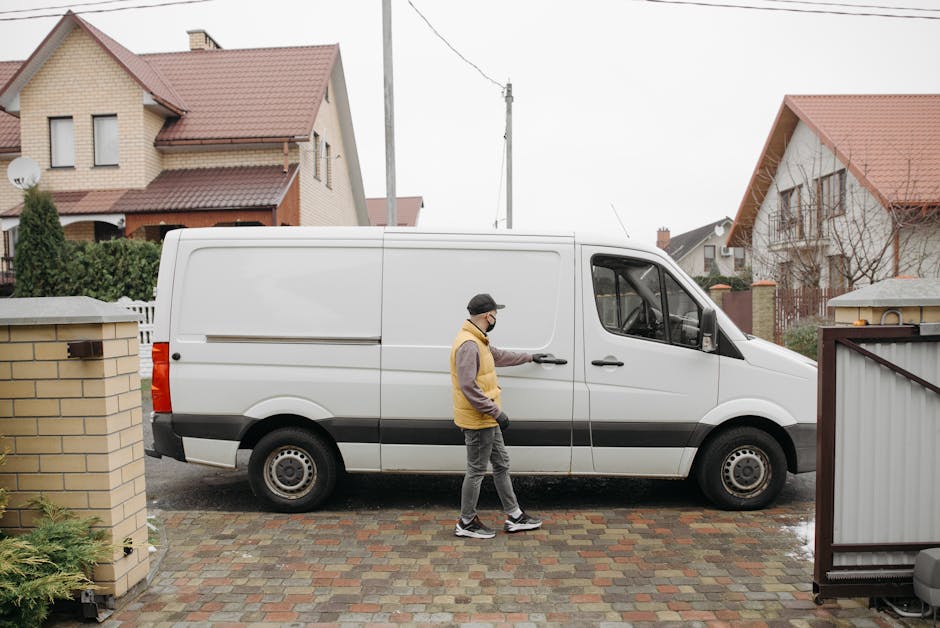 A man wearing a black cap, glasses, a beige and brown jacket, and gray trousers is standing beside a white commercial van, opening the sliding side door with his right hand during a home relocation process. The van is parked on a brick driveway adjacent to a residential property with a brick wall and a gate in Waddon. The driveway includes cobblestone paving, and there are some puddles indicating recent rain. In the background, there are detached houses with sloped roofs covered in red tiles, some with dormer windows and small front gardens. The setting is outdoors, under an overcast sky, with visible power lines overhead. The scene depicts the loading or unloading phase of furniture transport and packing materials, such as cardboard boxes and plastic wrap, as part of the house removal service offered by Man with Van Waddon, aligning with the topic of comparing self-hire versus professional removal options.