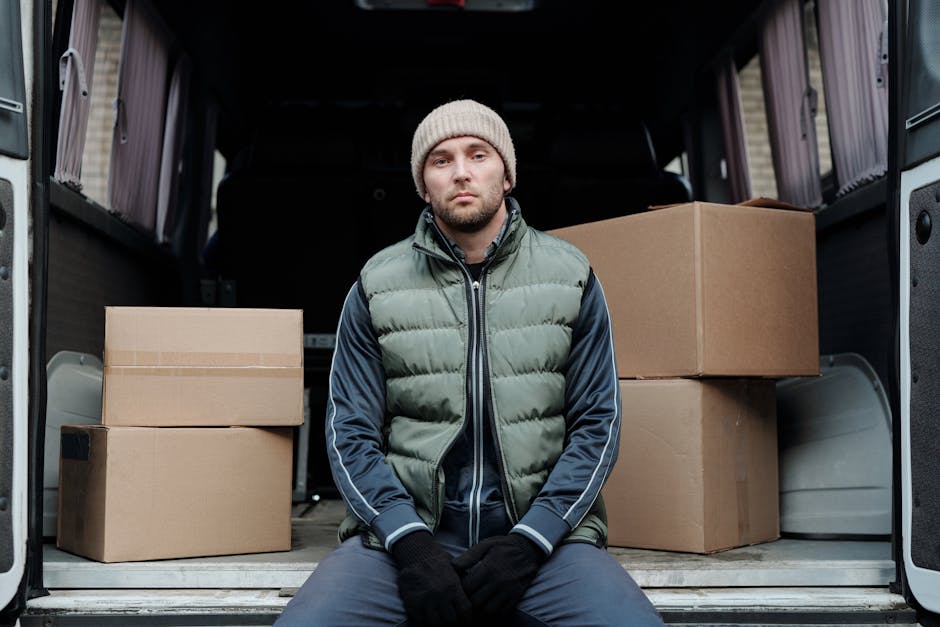 A young man with a beard, wearing a red baseball cap, a brown and beige jacket, a checked shirt, and striped gloves, sits on the rear bumper of a van with its back doors open during daylight. Inside the van, several cardboard boxes of varying sizes are arranged, with some stacked on top of each other, along with packaging materials. The interior of the van is illuminated by natural light, revealing a clean, organized space used for home relocation or furniture transport. The boxes are sealed with packing tape, and the floor of the van is visible in the foreground. The setting suggests a moving or packing process typical of professional removals, with the placement of furniture and boxes indicating the loading or unloading stage of a house move. This image illustrates aspects of packing, loading, and transport involved in services like those offered by Man with Van Waddon, fitting into the context of house removals and moving logistics.