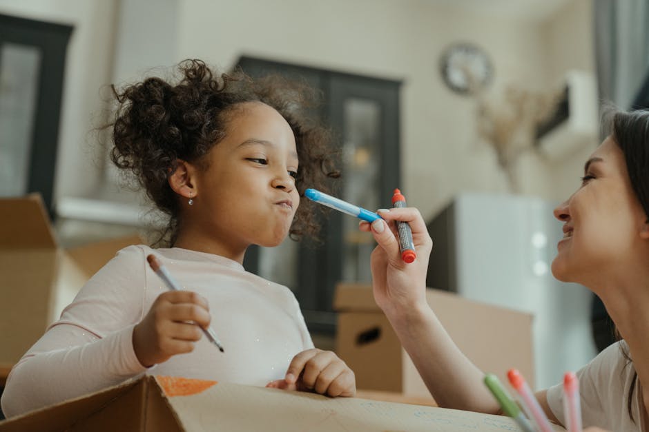 A young girl with curly hair, wearing a light-colored long-sleeve shirt, is inside a home surrounded by cardboard boxes, indicating a household packing or moving process. She is holding a pen in her right hand and appears to be making marks or taking notes on a box in front of her, while a woman with dark hair on the right, smiling, is gently holding a blue and red marker close to the girl's face, possibly helping or encouraging her. The room has neutral-toned furniture, including a dark cabinet with glass doors and a white appliance, with natural light coming through a window partially covered by curtains. The scene captures a moment of packing or home relocation, with the woman supporting the child's activity, consistent with furniture transport and packing steps often involved in house removals, as offered by Man with Van Waddon.