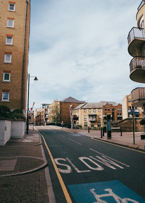 A street scene featuring a paved road in an urban residential area with multi-storey apartment buildings and houses on both sides. The road has a prominent white 'SLOW' marking and a blue parking space indicator near the curb. On the left, there is a tall street lamp, and on the right, balconies extend from a modern building. The area appears calm with minimal traffic and a single pedestrian visible in the distance. The environment is well-lit with natural daylight, under a partly cloudy sky. This setting exemplifies a typical urban street where home relocation and furniture transport services, such as those provided by Man with Van Waddon, might operate during a house moving process involving loading or unloading on the street or within close proximity to the property.