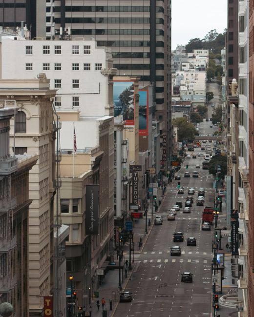 A high-angle view of a busy city street during daytime, showing multi-storey office and commercial buildings on either side with ornate facades and modern glass structures further down the road. The street is populated with numerous vehicles including cars, buses, and taxis, some waiting at traffic signals, with additional vehicles visible in the distance. Pedestrians walk along the sidewalks, which are lined with street lamps, signage, and shopfronts. In the foreground, the street's pavement is marked with crosswalk lines, and traffic lights are present at intersections. The scene captures an active urban environment, representing a typical city centre area suitable for services such as home relocation and furniture transport, with buildings and streets maintained in good condition. The natural daylight highlights the varied architectural styles and bustling city life, aligning with the moving and removals process managed by companies like Man with Van Waddon.