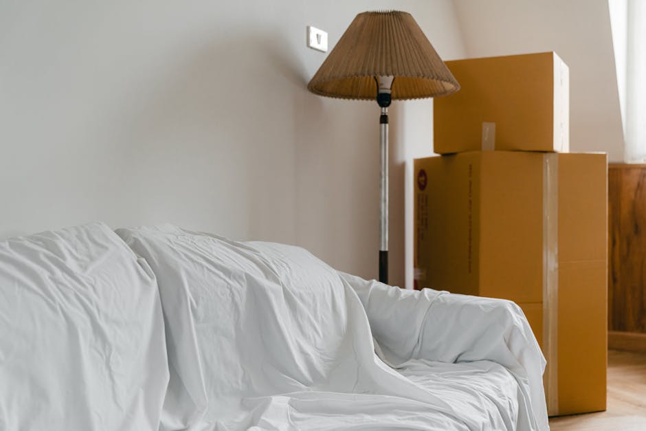 Inside a room with beige walls and wooden flooring, several cardboard boxes of varying sizes are stacked next to a wooden cabinet. The boxes are sealed with packing tape and appear to be prepared for
