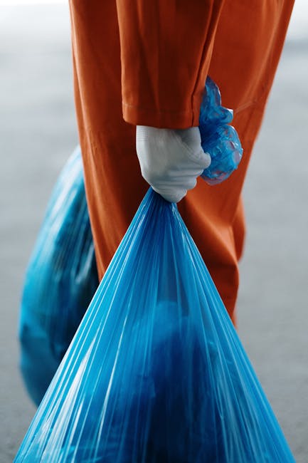 A person wearing orange overalls and white gloves is holding a large blue plastic bag filled with waste or bulky materials, suggesting waste collection or disposal. The individual is standing outdoors, with the background blurred, indicating an industrial or waste management environment. This scene relates to house removals and clearance services offered by Man with Van Waddon, especially when disposing of large items or bulky waste during a home relocation or clearance process. The focus is on the hand gripping the bag tightly, emphasizing proper handling and safety during waste collection, which complements professional moving and packing services. The image visually supports the process of removing bulky waste, in line with council rules and regulations, and highlights the importance of safe handling during household relocations.