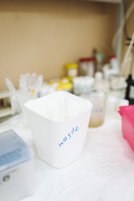 A white cardboard waste bin labeled 'waste' in blue handwriting is positioned on a white surface indoors, surrounded by various plastic containers, bottles, and jars filled with liquids, likely related to packing or cleaning materials. In the background, shelves hold numerous bottles and containers of different sizes and colors, including yellow, white, and red caps, indicating storage of household or cleaning supplies. The scene appears to be part of a packing or preparation area during a home relocation process managed by Man with Van Waddon, with the focus on organizing waste materials before moving. The lighting is neutral, illuminating the workspace clearly, reflecting a professional environment involved in house removals and furniture transport services, emphasizing efficient packing and waste management prior to transportation.