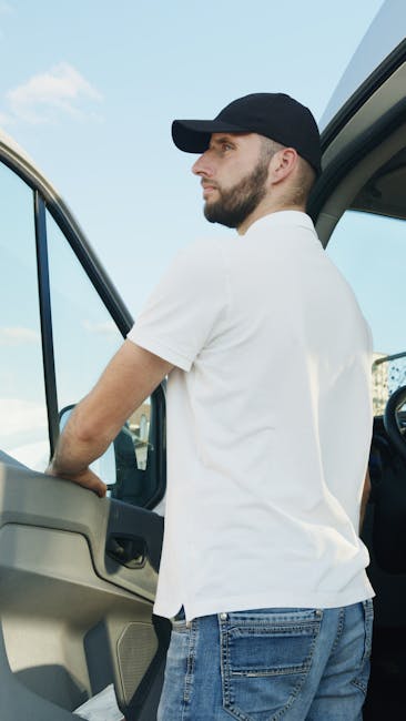 A man wearing a black cap and white t-shirt is standing outside the cab of a light-colored van, likely a moving vehicle used for house removals, with his arms resting on the door. The van is parked outdoors on a paved area under a clear blue sky, with a glimpse of a building in the background. Inside the van’s open side door, visible items include several cardboard boxes, some wrapped in plastic or fabric, and protective blankets used for furniture transport. The scene reflects the loading process typical of home relocation services provided by Man with Van Waddon, with an emphasis on efficient packing, lifting, and furniture transport. The grey dashboard and steering wheel can be seen inside the vehicle, indicating the vehicle is ready for loading or unloading of household goods, supporting efficient and careful packing and moving operations.