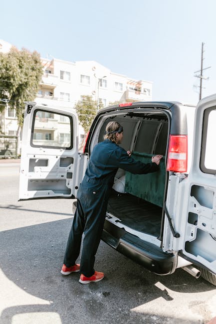 A person wearing a dark blue jumpsuit and red shoes loading a large green object into the open rear cargo area of a white van on a street in Waddon. The van is part of a home relocation or furniture transport service provided by Man with Van Waddon, with the rear doors swung open to facilitate the loading process. The vehicle is parked on the pavement near a row of residential buildings with white facades and balconies. In the background, there are trees, utility poles, and a clear sky, indicating a bright daytime setting. The interior of the van's cargo space is visible, showing a clean, empty area ready for a household item, and the individual is carefully maneuvering the object to ensure it fits securely inside during packing and moving activities characteristic of professional removals services.