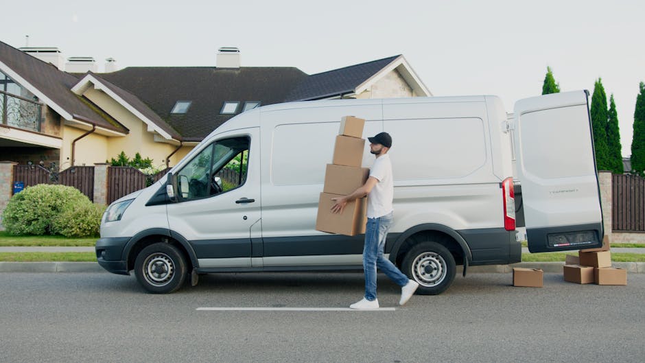 A man wearing a black cap and white t-shirt is standing outside the cab of a light-colored van, likely a moving vehicle used for house removals, with his arms resting on the door. The van is parked outdoors on a paved area under a clear blue sky, with a glimpse of a building in the background. Inside the van’s open side door, visible items include several cardboard boxes, some wrapped in plastic or fabric, and protective blankets used for furniture transport. The scene reflects the loading process typical of home relocation services provided by Man with Van Waddon, with an emphasis on efficient packing, lifting, and furniture transport. The grey dashboard and steering wheel can be seen inside the vehicle, indicating the vehicle is ready for loading or unloading of household goods, supporting efficient and careful packing and moving operations.