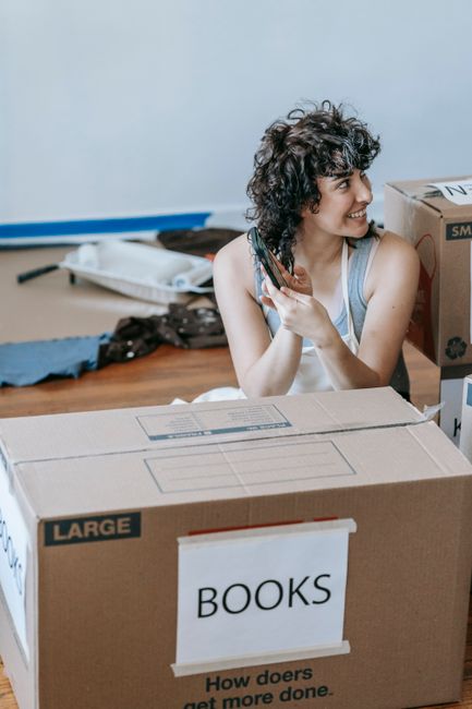 A young man and woman are inside a room filled with cardboard moving boxes, some labeled with items like 