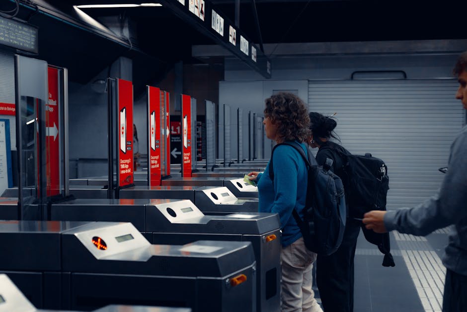 Three individuals are standing in front of self-service check-in kiosks at a train station, with the middle person operating a station touchscreen device while wearing casual clothing and backpacks. The environment features a row of kiosk machines lined up along a wall, with red signage indicating instructions for ticketing or transportation services. The lighting is bright, highlighting the modern, utilitarian design of the station area. The scene captures an aspect of travel planning, often related to home relocation or transportation logistics, which can be relevant for moving services. The individuals appear to be engaged in checking or buying tickets, with no luggage or heavy furniture visible, but the setting suggests the preparatory phase of a journey connected to house removals or furniture transport, as handled by companies like Man with Van Waddon.