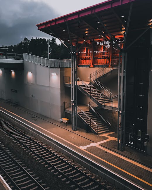 A view of the exterior of Waddon Station during dusk, showing a modern, multi-level entrance with metal stairs and handrails leading up to the station's upper walkways. The station building features large glass windows and is illuminated by outdoor lighting, casting a warm glow against the darkening sky. The station platform, situated alongside the railway tracks with visible sleepers and rails, is empty. The surrounding environment includes nearby trees and street lamps, with the scene capturing the calm atmosphere of a typical urban train station. As part of home relocation and furniture transport services, Man with Van Waddon utilises such stations for efficient access during tight-access moving jobs, coordinating loading and unloading that involve careful handling of boxes, furniture, and moving equipment near the station premises.