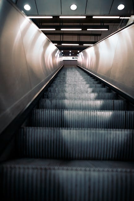 Three individuals are standing in front of self-service check-in kiosks at a train station, with the middle person operating a station touchscreen device while wearing casual clothing and backpacks. The environment features a row of kiosk machines lined up along a wall, with red signage indicating instructions for ticketing or transportation services. The lighting is bright, highlighting the modern, utilitarian design of the station area. The scene captures an aspect of travel planning, often related to home relocation or transportation logistics, which can be relevant for moving services. The individuals appear to be engaged in checking or buying tickets, with no luggage or heavy furniture visible, but the setting suggests the preparatory phase of a journey connected to house removals or furniture transport, as handled by companies like Man with Van Waddon.